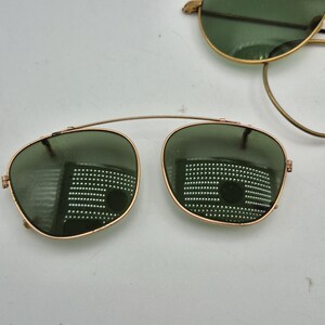 May include: Gold-rimmed sunglasses with green lenses. The sunglasses have a classic aviator style with a thin gold bridge connecting the two lenses. The lenses are oval-shaped and reflect the surrounding environment. Another pair of sunglasses is visible in the background.
