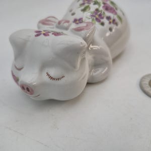 Puede incluir: Figura de cerdo de cerámica blanca en posición de dormir, adornada con motivos florales rosas y un lazo. El cerdo tiene los ojos cerrados y un hocico rosa. Una moneda está al lado de la figura.
