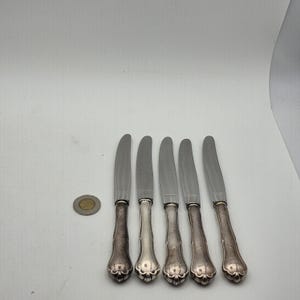 May include: Five silver-toned metal knives with curved blades and ornate, textured handles arranged in a row next to a small, round, two-toned metallic coin. The knives feature a polished finish on the blades and a darker, aged patina on the handles.
