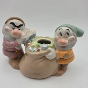 May include: A ceramic figurine featuring two characters with large noses and hats, one wearing a mauve tunic and the other an orange tunic, both holding a central tan sack filled with colorful, faceted gemstone-like shapes.