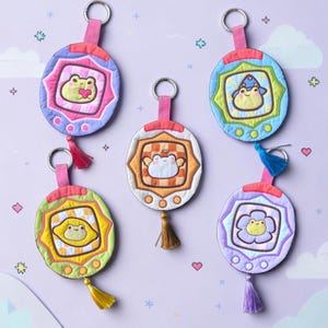 Puede incluir: Cinco llaveros de tela circulares con ranas bordadas en varios atuendos, cada uno con un anillo de metal, una trabilla de tela y una borla colgante. Los diseños incluyen una rana con un corazón, un mago, un limón, un ángel y una flor.