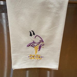 Könnte beinhalten: Cremefarbenes Küchentuch mit einem gestickten Wikinger-Logo in Lila, Gold und Schwarz. Das Wort "SKOL" ist darunter in goldener Schrift gestickt. Das Handtuch hängt an einer Metallstange.