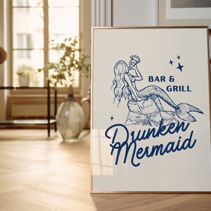 May include: A white sign with a gold frame featuring a blue illustration of a mermaid holding a drink. The text on the sign reads "Bar & Grill Drunken Mermaid" with stars on either side of the text.