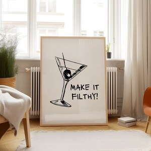 May include: A black and white illustration of a martini glass with an olive, with the text "Make it Filthy!" printed below the glass. The illustration is on a white background and framed in a light wood frame.