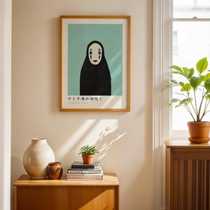 a framed poster of a black figure with a white face, a white vase, a stack of books, and a potted plant on a wooden table.
