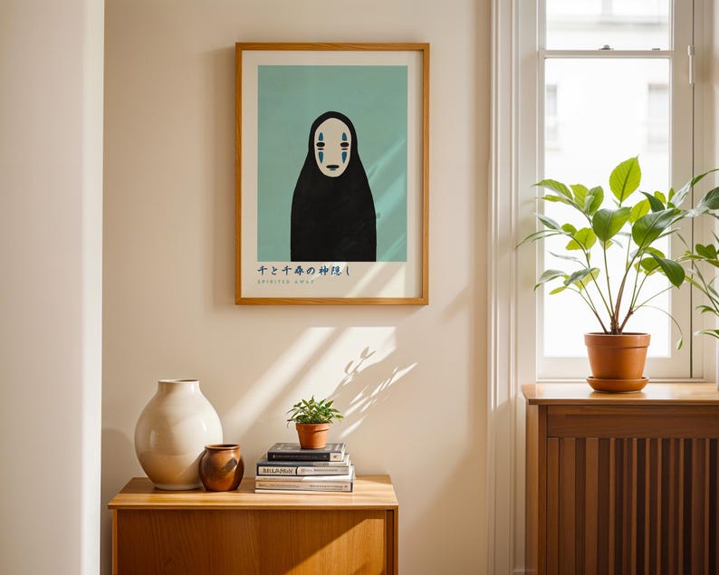 a framed poster of a black figure with a white face, a white vase, a stack of books, and a potted plant on a wooden table.