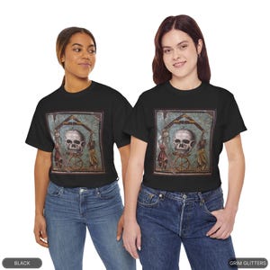 May include: Two people wearing black t-shirts with a graphic print of a skull in a frame. The skull is surrounded by figures and a wheel. The text "GRIM GLITTERS" is printed on the bottom right corner of the image.