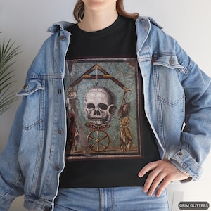 May include: A black t-shirt with a vintage-style image of a skull with a wheel and other symbols. The image is surrounded by a blue and green mosaic-like frame.