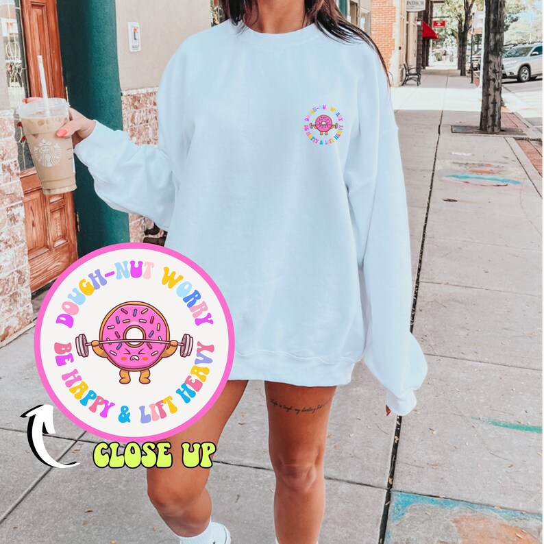 May include: A white sweatshirt with a pink circle graphic featuring a donut with a barbell and the text "Dough-Nut Worry Be Happy & Lift Heavy".
