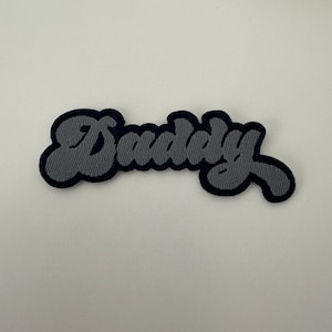 Könnte beinhalten: Ein grauer gestickter Aufnäher mit dem Wort "Dudley" in einer stilisierten Schrift.