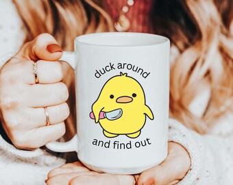 May include: White ceramic mug with a yellow cartoon duck holding a knife. The mug has the text "duck around and find out" printed on it.