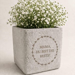 Könnte beinhalten: Ein quadratischer, grauer Beton-Blumentopf gefüllt mit weißen Blumen. Der Topf trägt den Text "MAMA, DU BIST DIE BESTE!" in einem kreisförmigen Design. Die Blumen sind klein und dicht, was ein volles, strukturiertes Aussehen erzeugt.