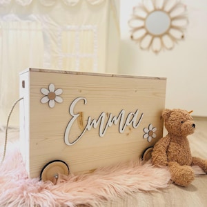 Könnte beinhalten: Eine hellfarbene Holzkiste mit dem Namen "Emma" in Schreibschrift, verziert mit Gänseblümchenmotiven. Ein Teddybär sitzt neben der Kiste, die auf einem rosa, flauschigen Teppich steht. Die Kiste hat Räder.