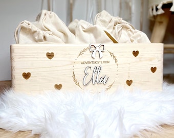 May include: A rectangular light-coloured wooden box featuring the text "ADVENTSKISTE VON Ella" in black, a white bow icon, and several small heart cutouts. The box is filled with multiple beige fabric drawstring bags.