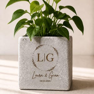 Könnte beinhalten: Ein quadratischer, hellgrauer Betonblumentopf mit einer grünen, belaubten Pflanze. Der Topf ist mit den Initialen "LIG" in einem kreisförmigen Design sowie den Namen "Laura & Gian" und dem Datum "08.05.2026" graviert.