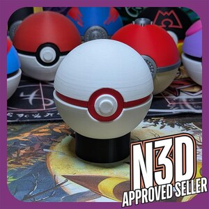 May include: A white Pokeball with a red ring and a small button in the center. The Pokeball is sitting on a black base. The background is a blurry image of a table with other Pokeballs.