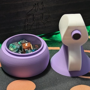 May include: Purple dice tower with a white top and a clear lid. The tower is open and contains several dice, including a green and blue iridescent die, a brown die with a 7 on it, and a black die with a 5 on it.