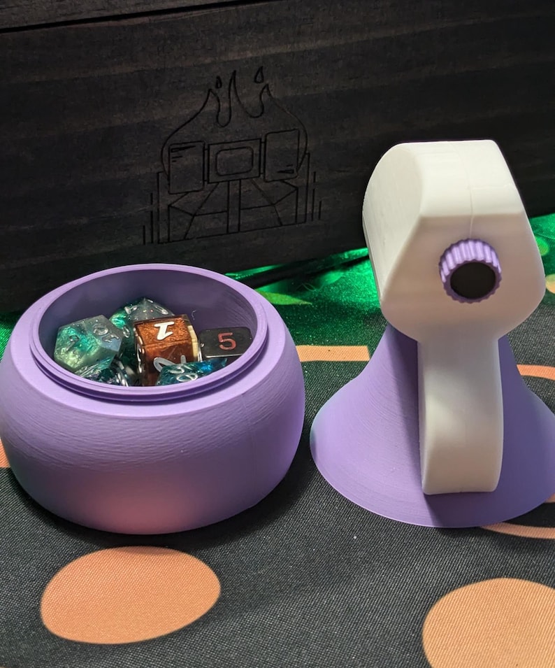 May include: Purple dice tower with a white top and a clear lid. The tower is open and contains several dice, including a green and blue iridescent die, a brown die with a 7 on it, and a black die with a 5 on it.