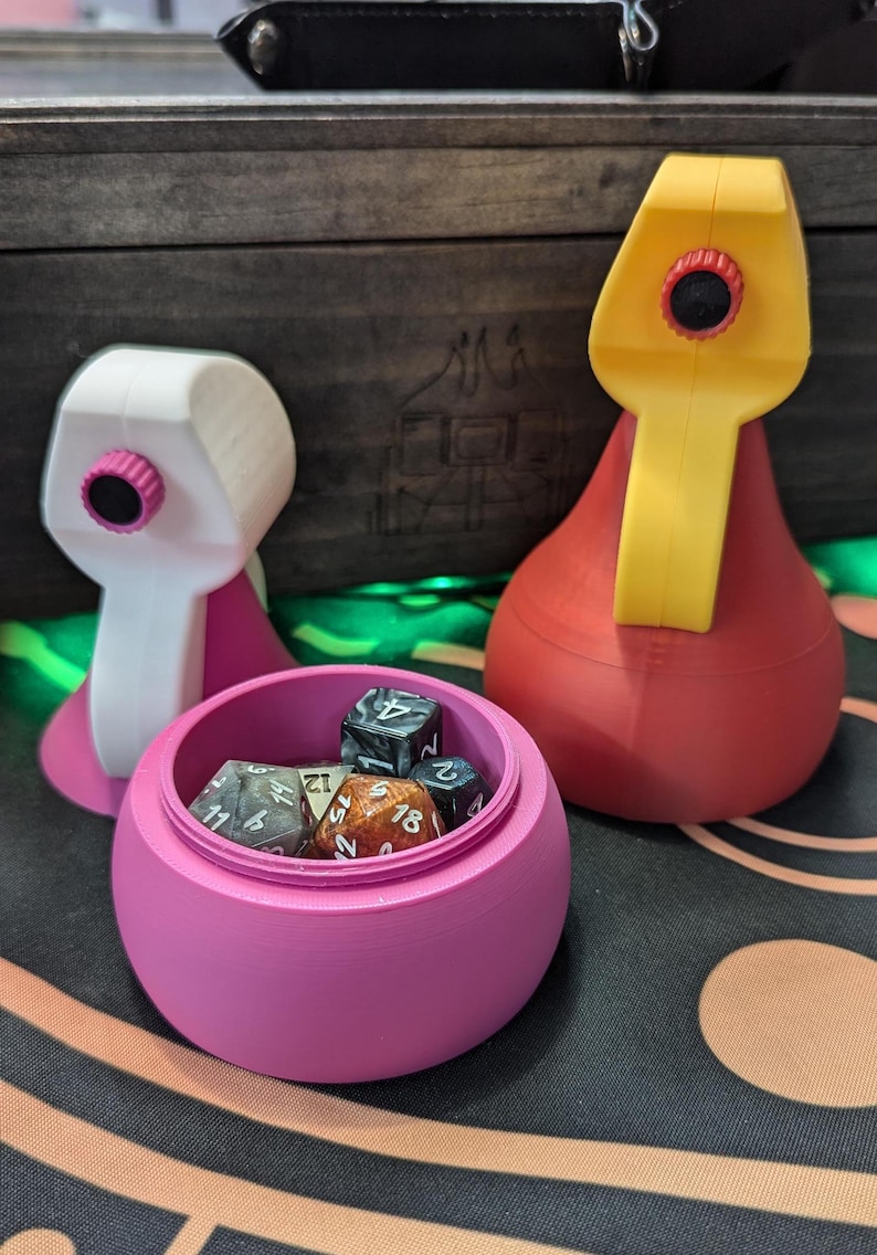May include: Two 3D printed dice towers, one white and one red, with a pink dice tower with a lid open and dice inside. The dice are various colors and have different numbers on them.