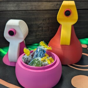 May include: A pink candy jar with a lid, filled with colorful wrapped candies. The jar is shaped like a rounded bowl with a small opening at the top. Two other colorful containers are visible in the background.
