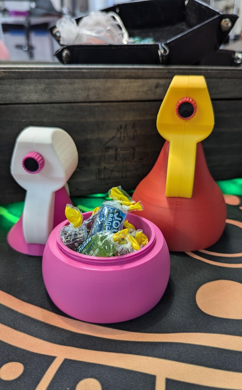 May include: A pink candy jar with a lid, filled with colorful wrapped candies. The jar is shaped like a rounded bowl with a small opening at the top. Two other colorful containers are visible in the background.