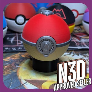 May include: A red, white, and gold Pokeball-style container with a silver latch. The container is sitting on a table with a black and white pattern.