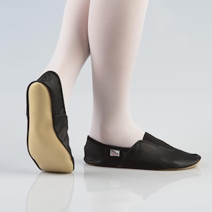 Op de afbeelding: Zwarte leren balletschoenen met een beige zool. De schoenen zijn ontworpen met een slip-on stijl en worden getoond op een persoon die een witte panty draagt. De schoenen hebben een klein label aan de zijkant.