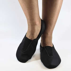 Op de afbeelding: Zwarte leren balletschoenen met een ronde neus en elastische bovenrand. De schoenen zijn ontworpen om nauwsluitend te zitten en zijn geschikt voor dans of andere activiteiten. De schoenen worden getoond op de voeten van een persoon tegen een witte achtergrond.
