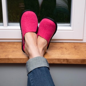 Pode incluir: Um par de chinelos de feltro rosa brilhante com acabamento preto. Os chinelos estão nos pés de uma pessoa, com as pernas apoiadas em uma superfície de madeira. A pessoa está usando jeans azul.