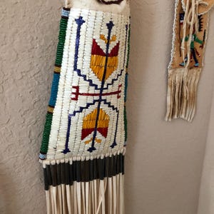 May include: A beaded Native American pouch with a fringe. The pouch is white with a geometric design in red, yellow, blue, and green. The pouch is hanging on a wall.