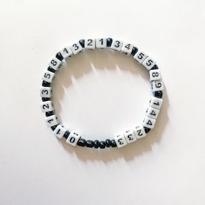 May include: A white and black beaded bracelet with number-printed cube beads. The bracelet features a repeating pattern of numbers from 0 to 9. The bracelet is made of small black beads and white cube beads.