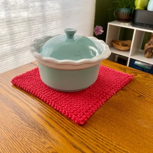 May include: A light blue ceramic casserole dish with a white rim and a lid sits on a red crocheted pot holder. The dish is on a wooden table.