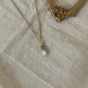 Pu&ograve; includere: Una collana a catena dorata con un piccolo ciondolo di perla e pietra preziosa verde.