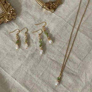 Pu&ograve; includere: Un set di orecchini d'oro e una collana con perline verdi e bianche. Gli orecchini sono orecchini pendenti con una perla in fondo. La collana &egrave; una semplice catena con una perla e perline verdi all'estremit&agrave;.