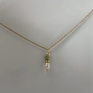Pu&ograve; includere: Una collana con catena dorata con un ciondolo con una perla bianca e tre pietre preziose verdi.