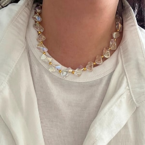 May include: A clear crystal necklace with gold-colored accents. The necklace features irregularly shaped, faceted crystals strung together with gold-colored beads. The necklace is worn over a white shirt.