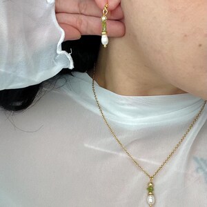 Pu&ograve; includere: Una collana a catena dorata con un ciondolo di pietra verde e perla. La collana &egrave; indossata da una persona con una camicia di rete bianca. La persona indossa anche un paio di orecchini dorati con pietre verdi e perle.