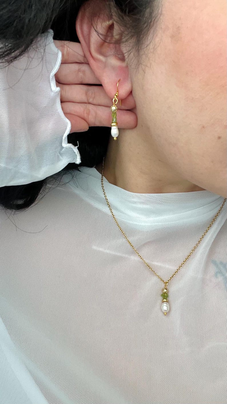 Pu&ograve; includere: Una collana a catena dorata con un ciondolo di pietra verde e perla. La collana &egrave; indossata da una persona con una camicia di rete bianca. La persona indossa anche un paio di orecchini dorati con pietre verdi e perle.