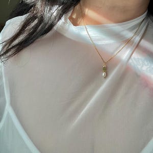 Pu&ograve; includere: Una delicata collana a catena dorata con un piccolo ciondolo di perla e pietra preziosa verde.
