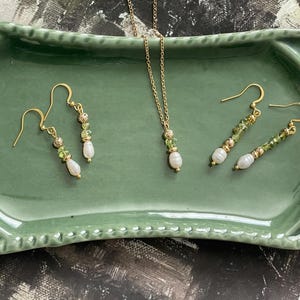 Pu&ograve; includere: Un set di collana e orecchini in oro con perline di pietre preziose verdi e un ciondolo di perla. I gioielli sono esposti su un vassoio in ceramica verde.