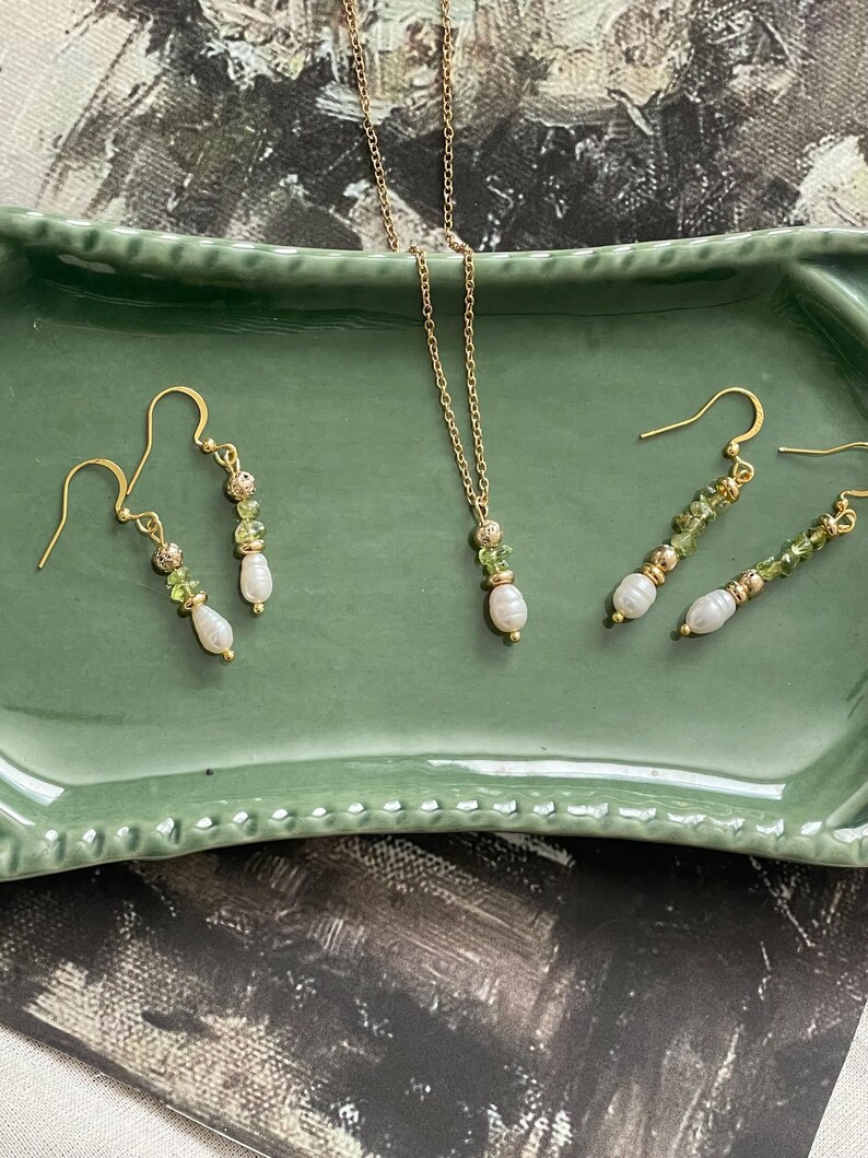 Pu&ograve; includere: Un set di collana e orecchini in oro con perline di pietre preziose verdi e un ciondolo di perla. I gioielli sono esposti su un vassoio in ceramica verde.