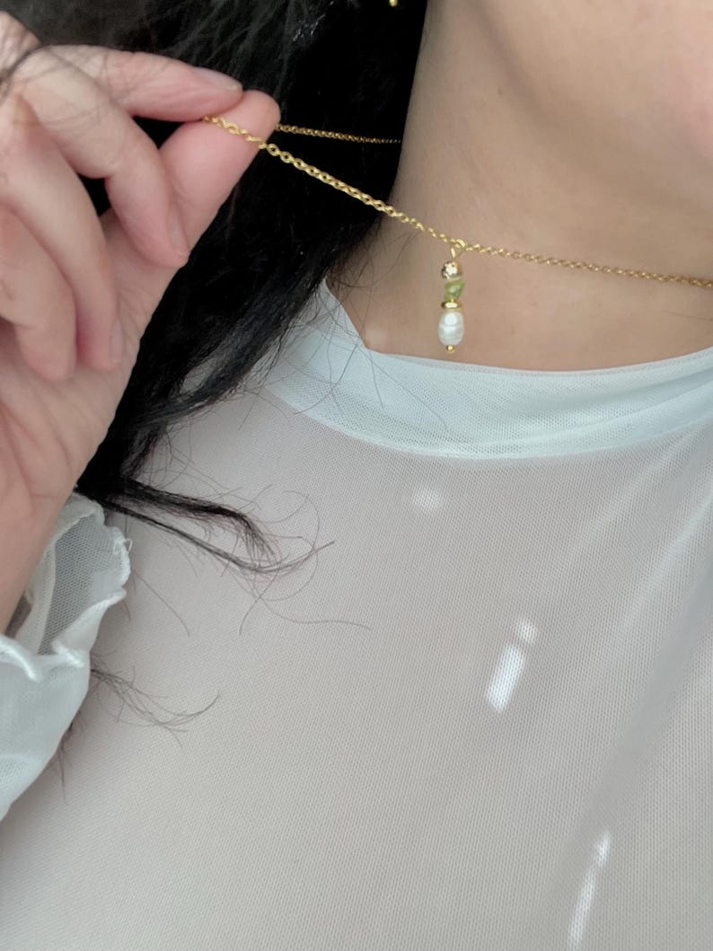 Pu&ograve; includere: Una collana a catena dorata con un piccolo ciondolo di perla e pietra preziosa verde.