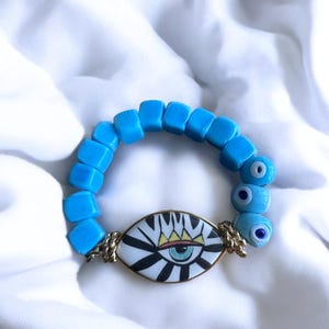May include: A blue and white evil eye bracelet with a large, colorful evil eye charm. The bracelet is made of square beads and has a gold chain connecting the charm to the beads.