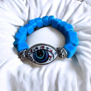 May include: A blue beaded bracelet with a silver charm featuring a white eye with blue, red, and black details.