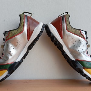 May include: A pair of silver, yellow, green, and red sneakers with black soles. The sneakers have a textured design and are set against a white background.