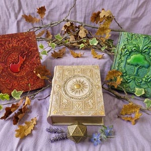 May include: Three embossed leather journals in red, cream, and green. The red journal has a flame, the cream journal a sun, and the green journal a tree. The journals are surrounded by dried leaves and sprigs, with a 20-sided die.