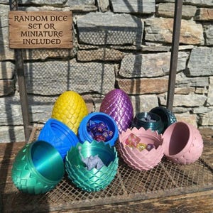 Può includere: Una collezione di contenitori per dadi colorati, con motivo a scaglie di drago, in vari colori, tra cui giallo, viola, blu, verde e rosa. Alcuni contenitori sono aperti, rivelando dadi o una miniatura. Un cartello di legno recita "RANDOM DICE SET OR MINIATURE INCLUDED."