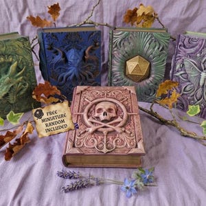 May include: A selection of fantasy books with detailed, embossed covers. Designs include a dragon, an octopus, a D20 die, and a butterfly. The central book features a skull and crossbones. A tag reads "FREE MINIATURE RANDOM INCLUDED". The books are arranged on a lilac surface.