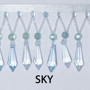 May include: A close-up of a light blue beaded trim with crystal-like teardrop-shaped beads. The trim has a white satin ribbon backing. The text "SKY" is printed below the trim.