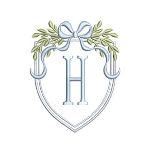 Op de afbeelding: Geborduurd monogramontwerp met de letter "H" in lichtblauw, geplaatst in een schildvorm. Een lichtblauwe strik en olijfgroene bladeren sieren de bovenkant, wat een elegant embleem creëert.